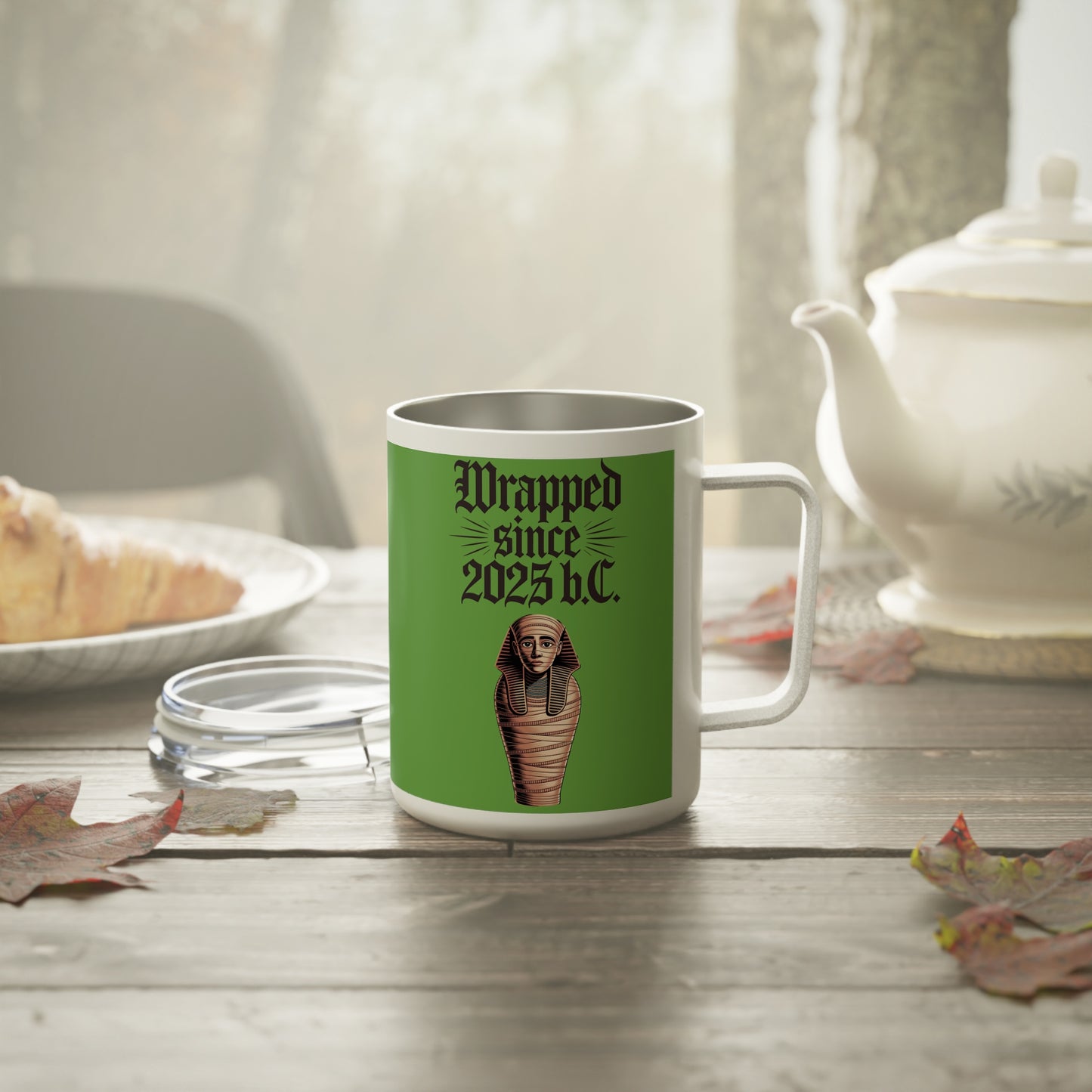 Wrapped Since 2025 B.C. stainless steel coffee mug on a table with autumn leaves and a teapot.
